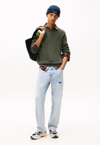 Oliven grønn genser over en rød og svart rutete skjorte, lyseblå slitte jeans, marineblå beanie og flerfargede sneakers. Svart totebag.