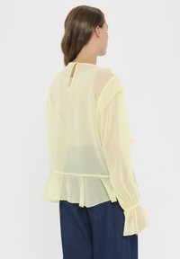 Gelbe, leichte Bluse mit Rüscheln an den Ärmeln und einem Peplum-Saum. Mit einem Schlüsselloch-Rücken und transparenter Textur. Kombiniert mit dunkelblauen Hosen.