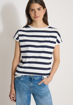 Femme aux cheveux foncés portant un t-shirt à manches courtes rayé blanc et marine et un jean bleu, debout avec une main dans la poche sur un fond uni.