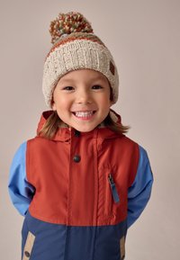 Színes, többrészes dzseki piros, kék és tengerészkék szakaszokkal; elöl cipzárral, patentos gombokkal és mellzsebbel rendelkezik. Texturált bézs kötött sapkával viselve, bojt díszítéssel.