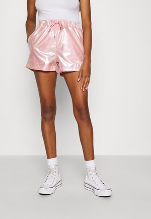 Personne portant un short rose brillant, un débardeur blanc, des chaussettes blanches et des baskets montantes blanches, debout, les mains reposant sur les côtés.