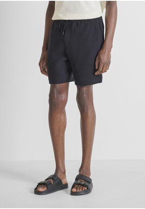 Mann trägt schwarze, strukturierte Shorts mit Kordelzug in der Taille und schwarze Sandalen mit Schnalle, stehend vor einem schlichten grauen Hintergrund.