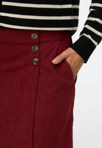 Jupe en velours côtelé rouge avec trois boutons marron à l'avant, des poches latérales et une surface texturée en relief.
