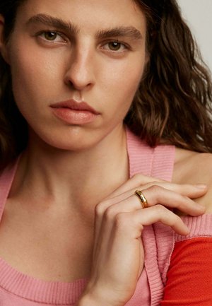 Anillo de oro en una mano con una superficie texturizada. La persona que lo lleva puesto viste un top de rayas rosa con acentos naranjas, mostrando tonos de piel naturales y cabello rizado.