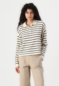 Long-sleeve polo sweater in cream with black horizontal stripes, featuring a collar and a loose fit. Paired with beige straight-leg pants.