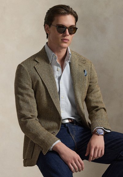 Νέος άνδρας με γυαλιά ηλίου, μπλέιζερ σε χρώμα μαυρίσματος με σχέδιο ψαροκόκκαλο, ριγέ πουκάμισο, σκούρο τζιν και ρολόι χειρός, καθισμένος μπροστά σε ουδέτερο φόντο.