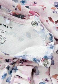 Tissu à motifs floraux dans des teintes pastel de rose et de violet, avec des boutons-pression en métal et des bords froncés doux pour une finition texturée.