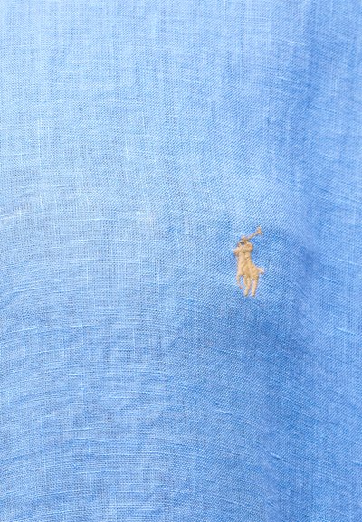 Šviesiai mėlynas tekstūruotas audinys su mažu kreminės spalvos siuvinėtu logotipu, vaizduojančiu žmogų, žaidžiantį polo ant žirgo.
