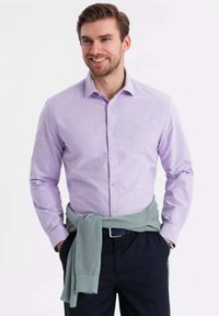 Camisa larga de color púrpura claro con cuello abotonado, combinada con un suéter de textura verde menta atado a la cintura, y pantalones oscuros.