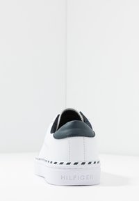 Baskets blanches en cuir lisse, accent en cuir bleu marine au talon, semelle extérieure rayée bleu marine et blanche, avec un logo en relief sur le côté.