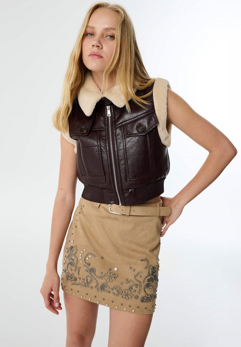 Chaleco corto de cuero marrón con un cuello de piel beige, combinado con una falda de ante color tierra que presenta adornos plateados y patrones florales.
