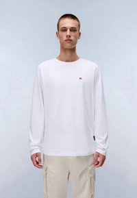 T-shirt branco de mangas longas com um pequeno logótipo vermelho no peito, apresentando um decote redondo e tecido de algodão suave. Combinado com calças de carga bege.