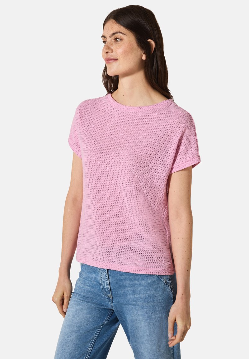 Pull over en tricot rose à manches courtes avec une coupe décontractée, présentant un motif en tissage ouvert texturé et un col rond. Porté avec un jean bleu clair.