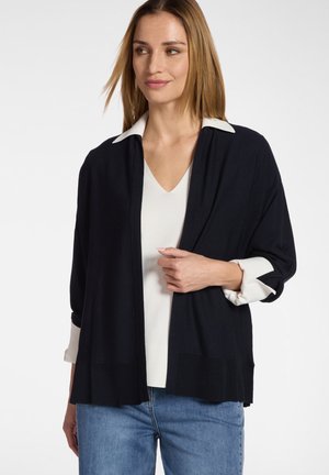 Vrouw met lichtbruin haar die een zwart vest met open voorkant draagt over een wit V-hals shirt en blauwe jeans, en lichtjes opzij kijkt.