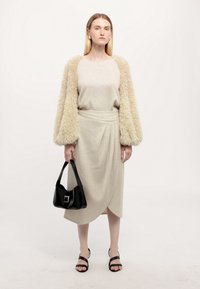 Cremfarbener, strukturierter Pullover mit flauschigen Ärmeln, kombiniert mit einem passenden, drapierten Rock. Das Model hält eine schwarze Umhängetasche und trägt schwarze Sandalen.