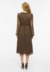 Brun prikkete kjole med lange ermer og volangkant på kragen. Gjennomsiktig stoff, innsvingt midje, midt på leggen, kombinert med svarte høye hæler.