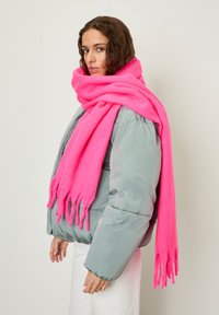 Woman wearing a bright pink oversized scarf, gray puffer jacket, and white pants, standing against a plain light background.