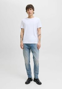 Camiseta blanca de algodón, jeans de mezclilla azul claros y zapatos negros de cordones. El modelo está de pie erguido frente a un fondo plano.