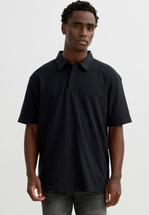 Joven con pelo corto y rizado, vestido con una camiseta polo negra lisa de manga corta y vaqueros oscuros, posando frente a un fondo claro y liso.