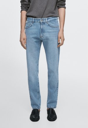 Person trägt hellblaue Jeans mit geradem Bein, dunkelgraues Langarmshirt und schwarze Lederloafers, steht vor einem einfarbigen Hintergrund.