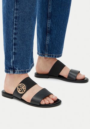 Schwarze Slipper-Sandalen mit zwei breiten Riemen, von denen einer eine dekorative, runde goldene Verzierung aufweist. Der Fuß ruht auf einer flachen, minimalistischen Sohle.