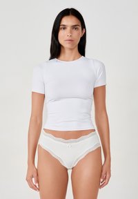 Top bianco a maniche corte in un tessuto liscio abbinato a biancheria intima chiara con bordi in pizzo e dettaglio a fiocco in vita.