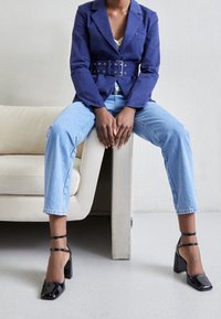 Blazer blu navy con cintura sopra un top chiaro, abbinato a jeans dritti blu chiaro e scarpe con tacco a blocco in vernice nera.