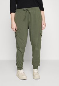 Pantaloni jogger verde oliva con vita elastica, dettagli con cordino e tasche laterali, abbinati a sneakers color crema.