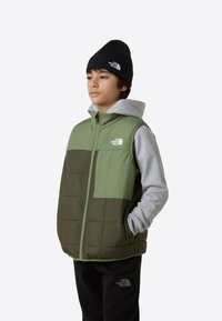 Grön och grå puffväst med dragkedja fram, med en bröstficka och kviltat mönster. Svart stickad mössa med logotyp. Svarta byxor.