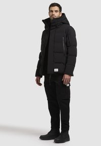 Jachetă puffer neagră cu guler înalt, buzunare cu fermoar și ajustări cu șnur. Asortată cu pantaloni cargo negri și cizme cu șireturi.