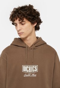 Brun hoodie i mjukt tyg med en känguru-ficka, prydd med en vit logotyp och dragsko för huvudet.