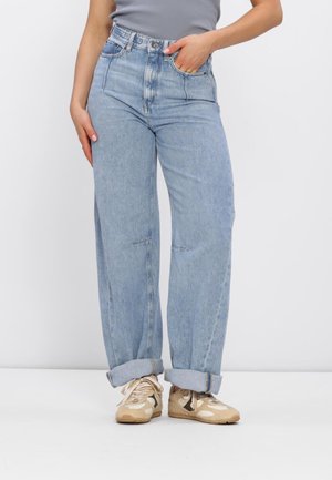 Person trägt hellblaue, umgeschlagene Jeans mit weitem Bein, graues Oberteil und beige Sneakers mit Schnürsenkeln, steht vor einem schlichten weißen Hintergrund.