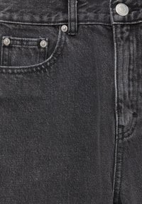 Gros plan sur un jean en denim noir montrant la poche avant, la petite poche avec des rivets en métal, la passants de ceinture et le bouton argenté sur la ceinture.