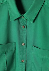 Chemise verte à col avec des boutons marron sur le devant et deux poches poitrine, montrant des coutures détaillées sur le tissu.