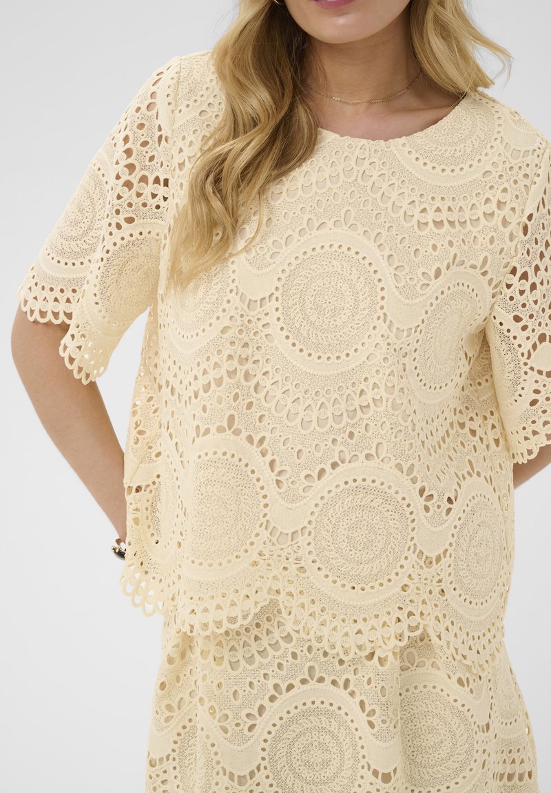 Femme blonde portant un haut en crochet crème et une jupe assortie avec des motifs circulaires et festonnés, les mains reposant sur les hanches.