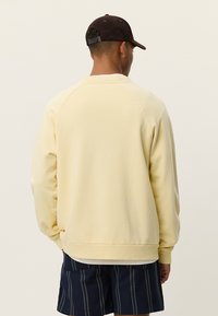 Άνδρας που φοράει ανοιχτό κίτρινο φούτερ, σκούρα μπλε ριγέ σορτς και μαύρο καπέλο μπέιζμπολ, με την πλάτη προς το μέρος μας, μπροστά σε απλό φόντο.