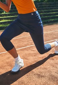Tengerészkék sportszoknya passzoló leggings felett, melynek derékpántján egy logó található, fehér sportcipőkkel párosítva, egy agyagpályán.