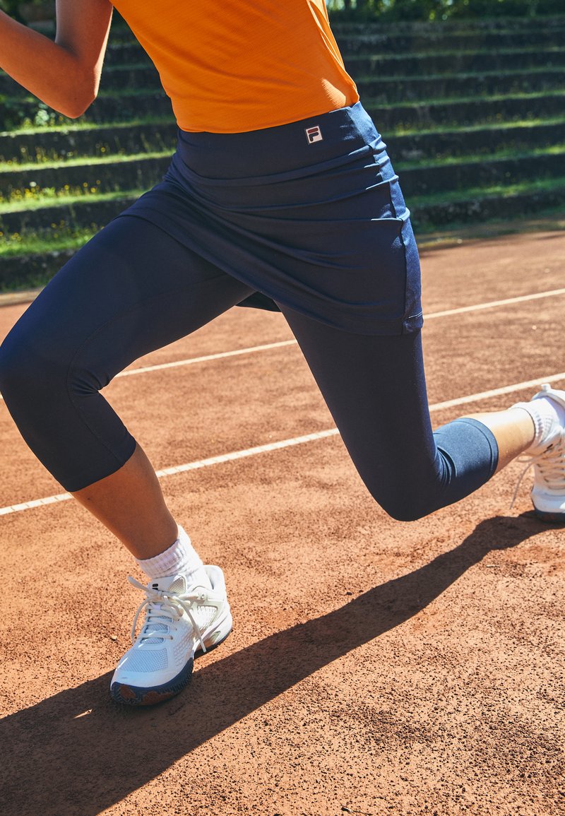 Tengerészkék sportszoknya passzoló leggings felett, melynek derékpántján egy logó található, fehér sportcipőkkel párosítva, egy agyagpályán.