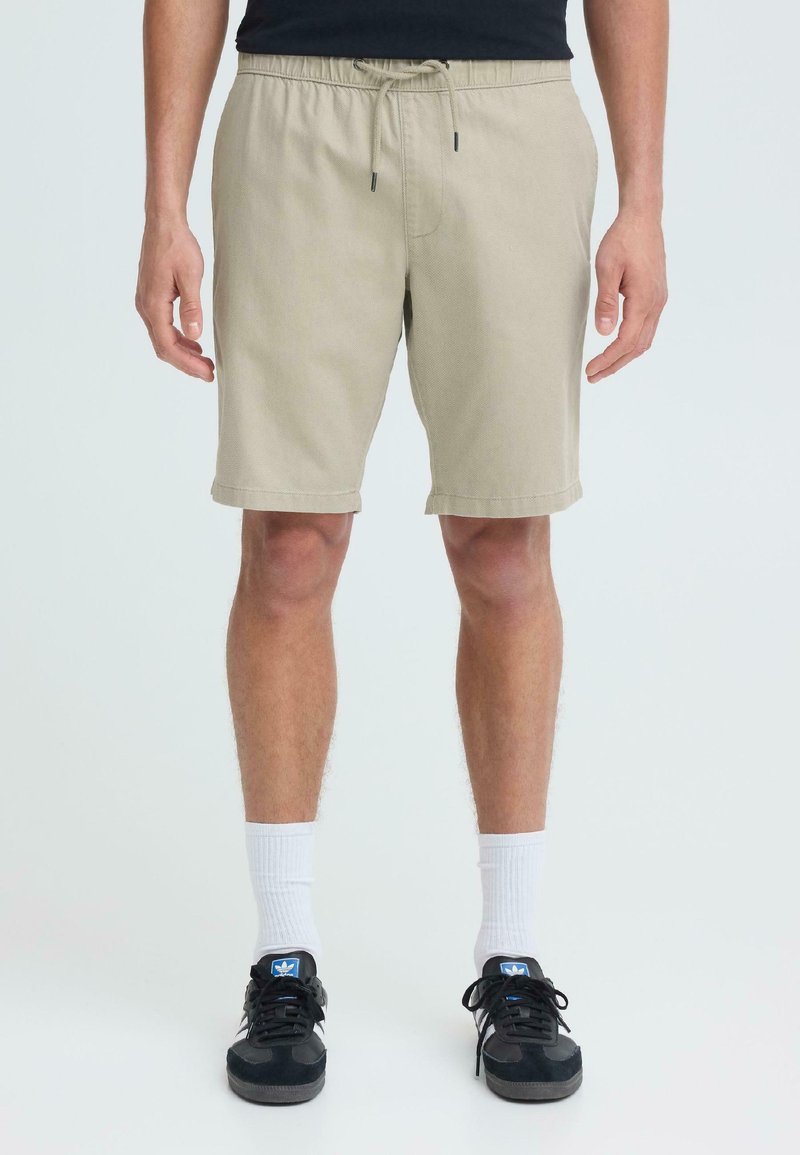 Homme portant un short beige à cordon de serrage, des chaussettes blanches mi-mollet et des baskets Adidas noires, debout devant un fond clair uni.