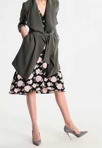 Frau in einem dunkelgrünen, taillierten Mantel über einem schwarzen Blumenkleid mit pinken Blumen, kombiniert mit grauen High Heels, steht auf einem Bein.