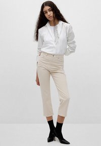 Beige enkellange broek met een strakke pasvorm, gecombineerd met een wit overhemd met knopen. Zwarte pumps met spits toe en enkellange zwarte sokken maken de look compleet.