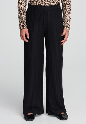 Persona che indossa pantaloni neri a coste a gamba larga, una maglietta a maniche lunghe con stampa leopardata e scarpe flat nere, in piedi contro uno sfondo semplice.
