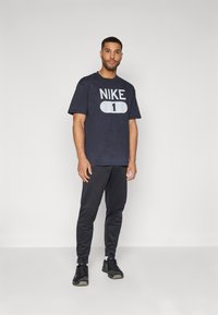 Marineblå Nike t-shirt med hvidt logo og nummer "1" foran, parret med sorte joggerbukser og sorte sneakers. Glat bomuldsstruktur.