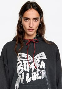 Melna kapuce, kas izgatavota no mīksta materiāla, ar lielu balto logo uzdruku un sarkanu šņori. Iekļauj priekšējo kabatu un pārāk lielu izmēru.