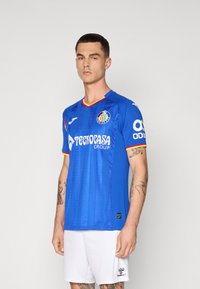 Modrá futbalová dres s krátkym rukávom, s červenými a žltými akcentmi, s logom na hrudi. Tkanina má hladkú, lesklú textúru.