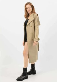 Beige trench coat s dvouřadým designem, velkými klopami a manžetami na opasek, zkombinovaný s černými kotníkovými botami s pevným podpatkem.
