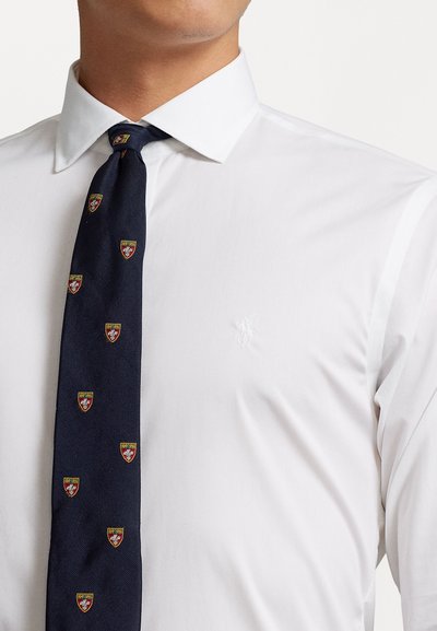 Chemise blanche à col classique, accompagnée d'une cravate bleu marine foncé ornée de petits motifs de boucliers brodés en rouge et jaune.