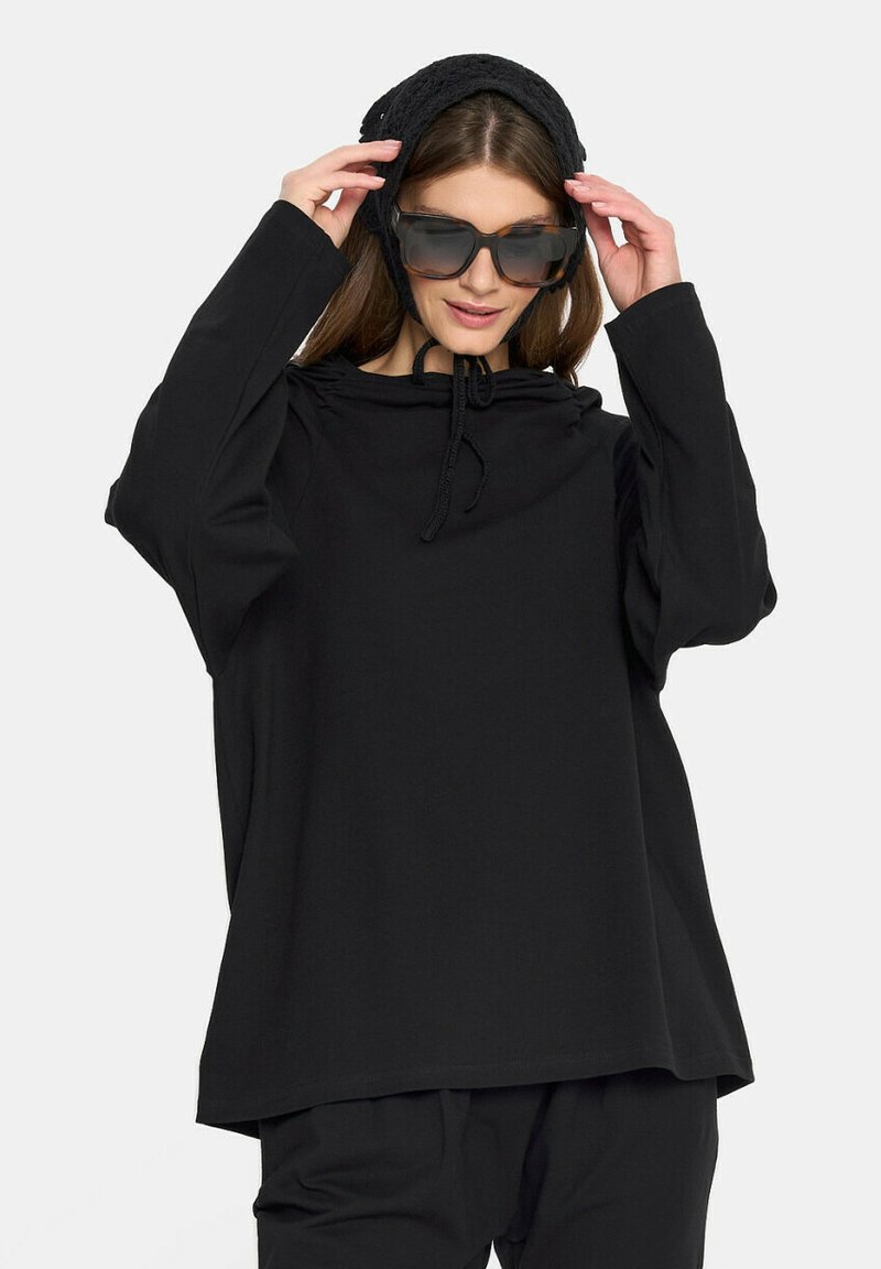 Czarny, oversize'owy sweatshirt z kapturem na sznurek, długimi rękawami i luźnym krojem; zestawiony z dużymi ciemnymi okularami przeciwsłonecznymi i czarną chustą na głowę.