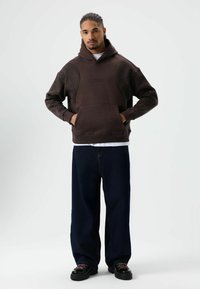 Bruine hoodie met patroonmouwen, kangoeroezak, geribbelde manchetten, gedragen met blauwe wijde jeans en zwarte schoenen. Textuuraccenten zichtbaar.