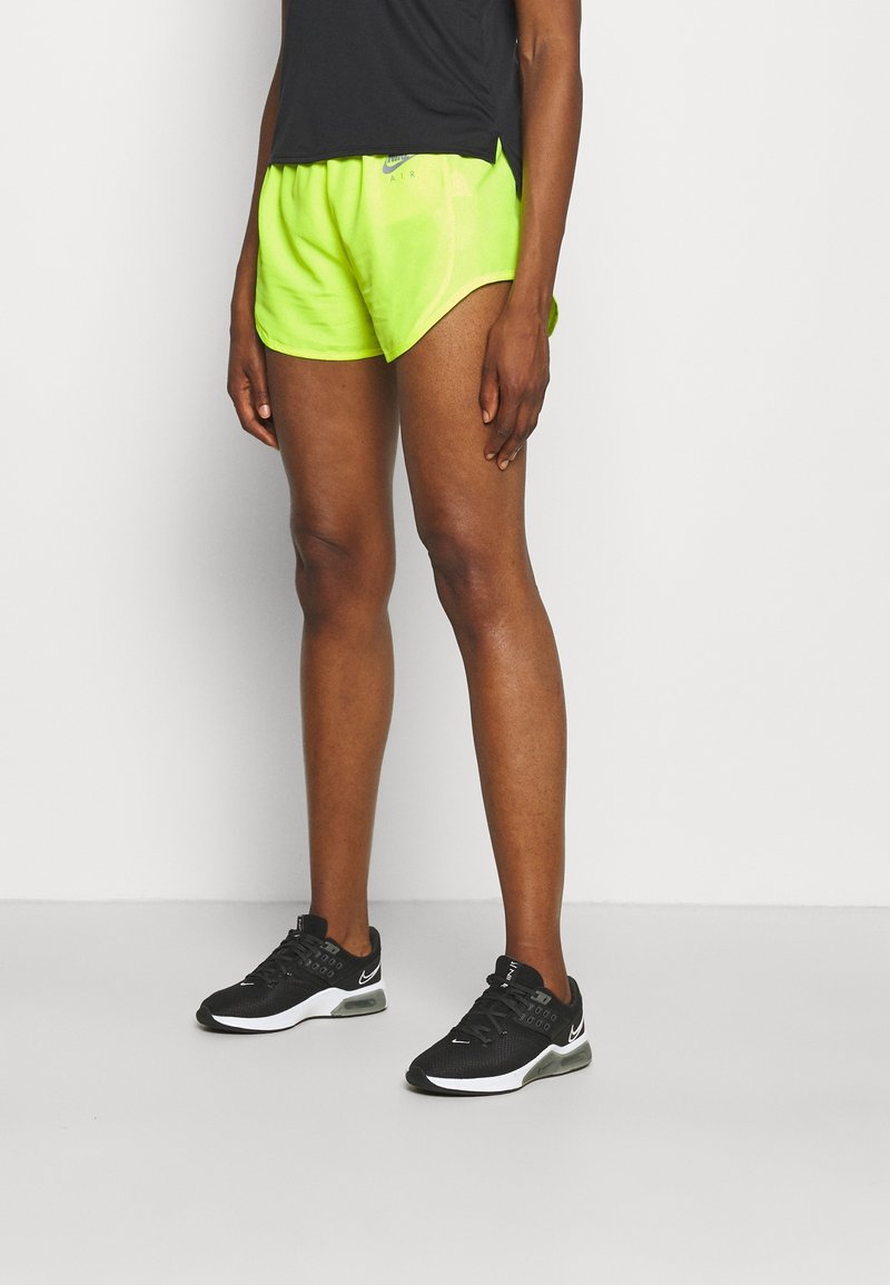 Pantalones cortos deportivos de neón verde con un dobladillo curvado, combinados con zapatillas negras que tienen suela blanca y parte superior de malla. Camiseta negra sencilla.